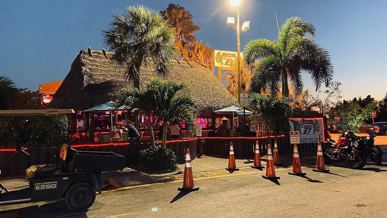Cafe 27 Bar & Grill tiki bar exterior at dusk with palm trees, thatched roof, motorcycles, and warm lighting in Southwest Ranches